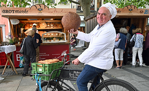 Unser Chef, Bäckermeister Ludwig Neulinger,  auf seinem Fahrrad hält einen großen Laib Brot vor dem Stand der Bäckerei Neulinger auf dem Brotmarkt hoch.